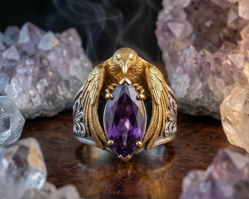 Puede incluir: Anillo de plata detallado con detalles dorados, con un dise&ntilde;o de ave que sostiene una gran piedra preciosa p&uacute;rpura con corte marquesa. El anillo se encuentra sobre un fondo de cristales de amatista.