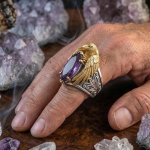 Puede incluir: Un anillo de oro y plata con un dise&ntilde;o de cuervo que sostiene una gran gema p&uacute;rpura de corte ovalado. El anillo tiene intrincados detalles de plumas y florales. El anillo se muestra en una mano, rodeado de cristales de amatista.