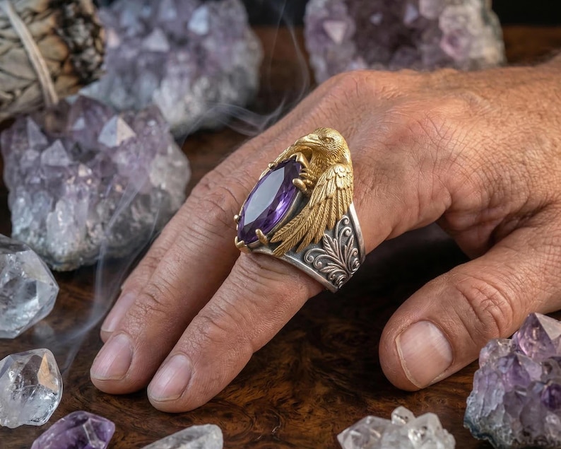 Puede incluir: Un anillo de oro y plata con un dise&ntilde;o de cuervo que sostiene una gran gema p&uacute;rpura de corte ovalado. El anillo tiene intrincados detalles de plumas y florales. El anillo se muestra en una mano, rodeado de cristales de amatista.