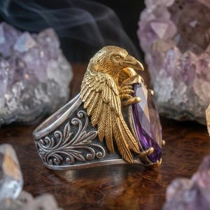 Puede incluir: Un anillo de plata detallado con un dise&ntilde;o de cuervo dorado que sostiene una gran gema p&uacute;rpura facetada. El anillo tiene intrincados patrones florales en la banda. Las alas del cuervo est&aacute;n extendidas y la gema es de color violeta intenso.
