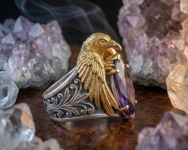 Puede incluir: Un anillo de plata detallado con un dise&ntilde;o de cuervo dorado que sostiene una gran gema p&uacute;rpura facetada. El anillo tiene intrincados patrones florales en la banda. Las alas del cuervo est&aacute;n extendidas y la gema es de color violeta intenso.