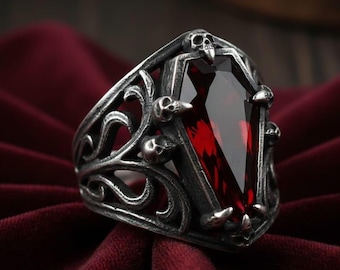 Puede incluir: Un anillo de tono plateado con una piedra preciosa de color rojo oscuro en forma de ataúd. La banda está decorada con volutas intrincadas y pequeños motivos de calaveras alrededor del engaste de la piedra, creando un diseño metálico detallado.