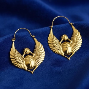 May include: Two gold-coloured hoop earrings with an Egyptian scarab beetle design. The earrings feature a winged scarab beetle pendant and are set against a deep blue fabric background. A unique piece of jewellery.