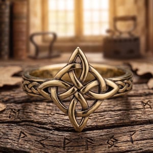Könnte beinhalten: Ein goldfarbener Ring mit keltischem Knoten und aufwendigem Design. Der Ring hat einen zentralen Knoten und ein geflochtenes Muster um das Band. Der Ring wird auf einer Holzoberfläche mit geschnitzten Symbolen präsentiert.