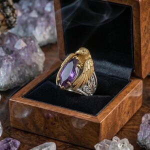 Puede incluir: Un anillo de oro y plata con un dise&ntilde;o de &aacute;guila, engastado con una gran gema p&uacute;rpura de corte ovalado. El anillo se exhibe en una caja de madera con interior de terciopelo negro, rodeado de cristales de amatista. Se destacan los intrincados detalles del anillo.