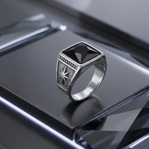 Op de afbeelding: Een zilverkleurige ring met een grote, vierkante, zwarte edelsteen. De ring heeft gedetailleerde zijontwerpen, waaronder een ster en een stippenpatroon. Het wordt gepresenteerd op een reflecterend, donker oppervlak.