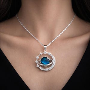 Peut inclure: Collier argenté avec un pendentif céleste. Le pendentif représente une planète bleue entourée d'un anneau, d'un croissant de lune et d'étoiles, tous ornés de cristaux transparents. Le collier est porté autour du cou d'une personne.