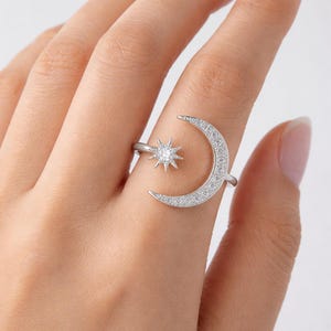 Può includere: Un anello color argento con un design a forma di luna crescente e stella. La luna è incastonata con piccole pietre trasparenti e la stella ha una pietra centrale più grande. Questo anello è a tema celeste.