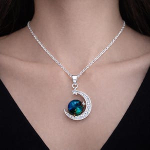 Puede incluir: Un collar plateado con un colgante de media luna engastado con cristales transparentes. La luna abraza un orbe azul y verde, con una pequeña estrella. El collar se lleva alrededor del cuello.