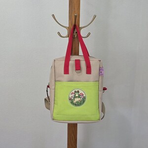 May include: A beige backpack with bright red top handles and a lime green front pocket. The pocket features a circular embroidered patch depicting a green frog sitting among red and white mushrooms and green foliage. The bag has a small metal D-ring.