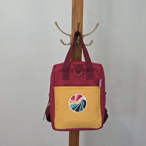 May include: A deep magenta backpack featuring a mustard yellow front pocket and two top handles. A circular patch on the front pocket displays a stylized sun rising over a dark blue wave. The bag includes a metal D-ring near the top handles.