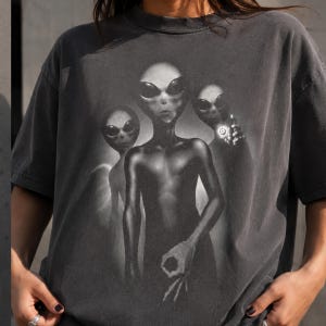 Op de afbeelding: Een antracietgrijs t-shirt met korte mouwen en een grafische print van drie buitenaardse wezens. De middelste alien maakt een handgebaar, terwijl de figuur aan de rechterkant een gloeiende bol vasthoudt. Het ontwerp is in grijstinten uitgevoerd.