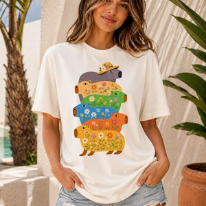 May include: An off-white short-sleeved t-shirt featuring a vertical stack of five capybara illustrations. Each capybara is a different colour, including grey, yellow, green, blue, and orange, and is decorated with colourful floral patterns.
