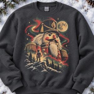 May include: A dark charcoal gray crewneck sweatshirt featuring a large graphic print of a nutcracker soldier with a white beard, a red uniform, and a tall hat. The design includes a full moon, swirling red and green aurora lights, and a small silhouette of a nutcracker standing on a snowy cliff surrounded by pine trees.
