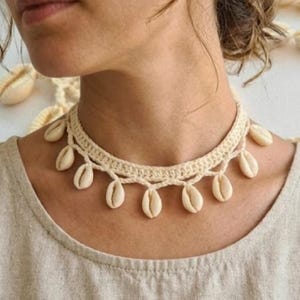 May include: Cream-colored crochet choker necklace adorned with multiple small seashells. The necklace is worn on a person's neck, and the shells hang down. The person is wearing a light beige top. The background is a light color.