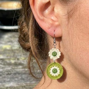 May include: Crocheted earrings with a floral design. The earrings feature a small white flower at the top and a larger green and white circular design below. The earrings are suspended from a silver hook and are shown against a blurred background.