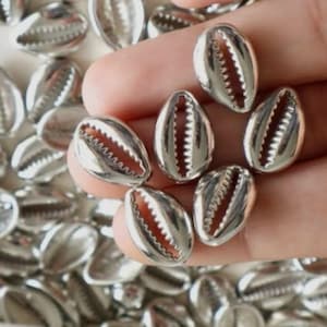 May include: A collection of small, metallic silver cowrie shell beads. Each bead features a smooth, reflective surface and a central longitudinal opening with serrated edges, mimicking the natural shape and texture of a sea shell.