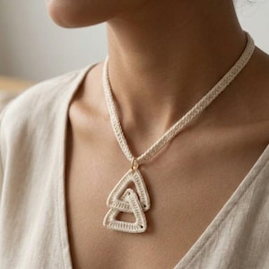 May include: Cream-colored crochet necklace featuring a pendant with two interlocking triangular shapes. The necklace has a braided cord and a gold-colored clasp. The pendant is made of the same crochet material as the necklace.