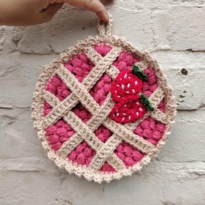 May include: A handmade crochet potholder shaped like a pie, featuring a lattice crust design and a strawberry applique. The potholder is primarily pink with tan and red accents, and has a loop for hanging.