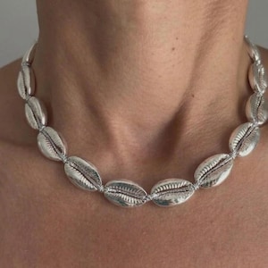 May include: A choker necklace featuring a series of metallic silver-toned cowrie shells strung together. The shells are uniform in size and shape, displaying a smooth, reflective surface with the characteristic central slit of a cowrie shell.
