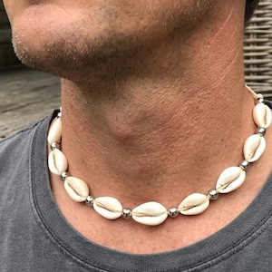 May include: A choker necklace featuring a series of white cowrie shells alternating with small, polished silver-toned metallic beads. The shells are strung together on a cord, creating a circular design that rests around the base of the neck.