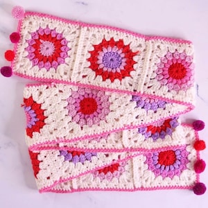 May include: A rectangular crochet scarf featuring a series of joined square motifs in cream, red, pink, and purple. The scarf is bordered with a bright pink yarn edge and decorated with small, colorful pom-poms attached along the short ends.