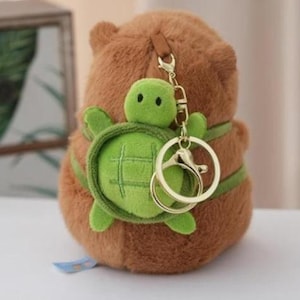 May include: A brown plush bear keychain with a green turtle attached. The turtle has a green shell and limbs. The keychain has a gold-colored clasp and ring. The bear is wearing a green belt.