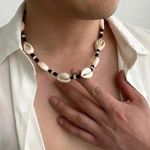 May include: A necklace featuring alternating white cowrie shells and small black cylindrical beads. The shells are arranged in a repeating pattern with the black beads, creating a contrast between the natural off-white tones and the dark beads.