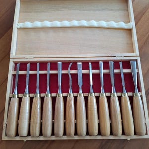 May include: A set of twelve wood carving chisels with light brown wooden handles and silver metal blades, arranged in a rectangular wooden storage box with a red velvet interior lining and a white foam strip on the inside of the lid.