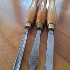 May include: Three wood carving chisels with light brown wooden handles and brass ferrules. The metal blades include a gouge, a spear point, and a flat chisel, with the text Stanley Bridges visible on the metal shafts of the two right tools.
