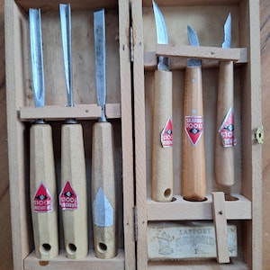 Op de afbeelding: Een set van zes houtsnijgereedschappen met lichtgekleurde houten handvatten en metalen bladen, opgeborgen in een rechthoekige houten doos. Elk handvat heeft een rood-wit Skandia Tools label. Een klein rechthoekig blok met het opschrift Lapport Record is inbegrepen.