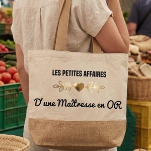 Peut inclure: Un sac fourre-tout en toile beige avec deux anses et une base en toile de jute marron. L'avant présente le texte noir LES PETITES AFFAIRES et D'une Maîtresse en OR, séparé par un cœur métallique doré avec des motifs de feuilles stylisées.
