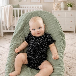 May include: A baby in a black polka dot onesie sits in a sage green textured baby seat, smiling. The baby has blue eyes and fair skin. A white cot and chest of drawers are in the background.