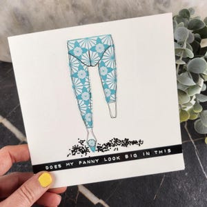Puede incluir: Una tarjeta de felicitación blanca con un borde negro en la parte inferior. La tarjeta presenta una ilustración de leggings con un estampado floral turquesa y blanco. El texto "DOES MY FANNY LOOK BIG IN THIS" está impreso en el borde negro.