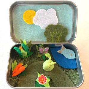 May include: A miniature felt scene inside a metal tin. The scene includes a yellow sun, white cloud, green tree, and a pond with a blue fish. Other elements include a green bunny, carrots, and a flower.
