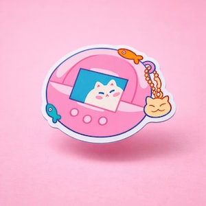 May include: A pink sticker featuring a retro handheld game design with a cat character on the screen. The sticker has a blue screen, three buttons, and a dangling charm with a cat face and fish. A small fish is on the left side.