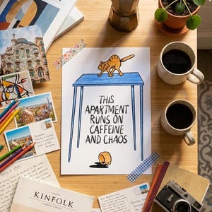 Puede incluir: Vista superior de una mesa de madera con varios objetos. En el centro, una impresión con el texto "THIS APARTMENT RUNS ON CAFFEINE AND CHAOS" y un gato de dibujos animados. Tazas de café, cafetera, postales y revistas también están presentes.
