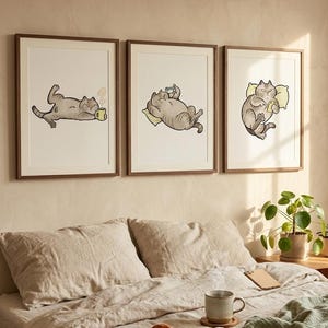 May include: Three framed art prints featuring cartoon cats in various relaxed poses. One cat lounges with a mug, another uses a phone, and the third rests on a pillow. The artwork has a neutral color palette and is displayed above a bed.
