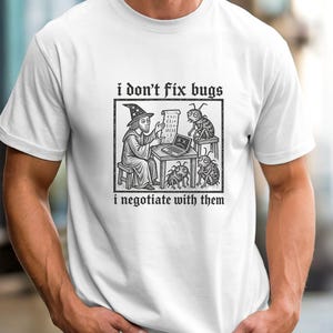 Op de afbeelding: Een wit t-shirt met korte mouwen en een zwarte grafische illustratie van een tovenaar die aan een bureau zit met een laptop en drie insecten. De tekst erboven luidt i don't fix bugs en de tekst eronder luidt i negotiate with them.