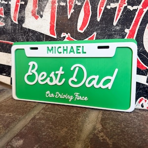 May include: A green license plate with the words "Best Dad" in white script. The name "Michael" is above the script, and "Our Driving Force" is below. The plate has a white border.