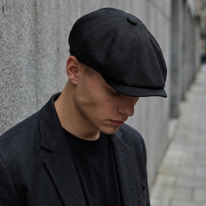 Può includere: Un cappello nero da ragazzo con una corona arrotondata e una piccola visiera. Il cappello è realizzato in tessuto strutturato e viene indossato con un blazer grigio scuro e una maglietta nera. Uno stile classico.