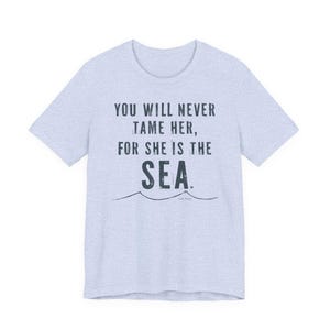 Puede incluir: Camiseta azul claro con la frase "YOU WILL NEVER TAME HER, FOR SHE IS THE SEA" en azul oscuro. Debajo del texto hay un dise&ntilde;o de olas.