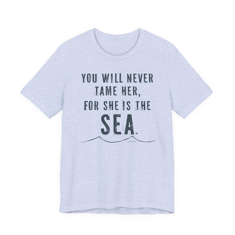 Puede incluir: Camiseta azul claro con la frase "YOU WILL NEVER TAME HER, FOR SHE IS THE SEA" en azul oscuro. Debajo del texto hay un dise&ntilde;o de olas.