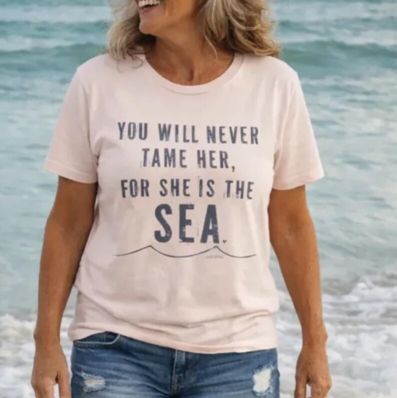 Puede incluir: Una camiseta rosa claro con el texto "YOU WILL NEVER TAME HER, FOR SHE IS THE SEA" en letras azul oscuro. La camiseta tiene cuello redondo y mangas cortas. El fondo muestra una escena oce&aacute;nica borrosa.