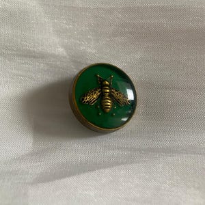 Pode incluir: Botão redondo verde com um desenho de abelha dourada. A abelha tem asas e corpo detalhados. O botão tem uma borda dourada e está sobre um fundo branco texturizado.