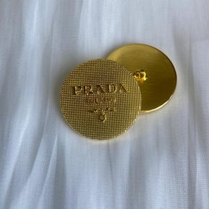 Pode incluir: Dois botões Prada dourados. Um botão exibe o nome e o logotipo da marca em relevo. O outro mostra a parte de trás do botão. Ambos os botões são redondos e têm uma superfície texturizada.