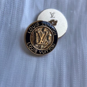 Può includere: Due bottoni rotondi in metallo dorato con il logo Louis Vuitton. Il bottone superiore presenta un bordo in smalto nero con la scritta Louis Vuitton impressa in oro, che circonda un centro dorato testurizzato con il monogramma LV. Il bottone posteriore è liscio.