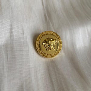 Op de afbeelding: Een goudkleurige knoop met een gedetailleerd Medusa-hoofdontwerp in het midden. De knoop heeft een Griekse sleutelrand en staat tegen een witte stoffen achtergrond. De knoop is rond en lijkt van metaal te zijn.