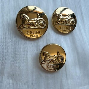 Könnte beinhalten: Drei runde, goldfarbene Metallknöpfe in verschiedenen Größen, jeweils mit einem geprägten Motiv einer Pferdekutsche und dem Schriftzug CELINE PARIS. Die Knöpfe haben eine polierte, reflektierende Oberfläche und eine kreisförmige Form.