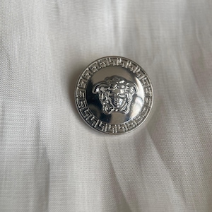 Puede incluir: Un botón redondo plateado con un diseño en relieve detallado. El botón presenta una imagen central de una cara rodeada por un borde decorativo. El botón está sobre un fondo de tela blanca.